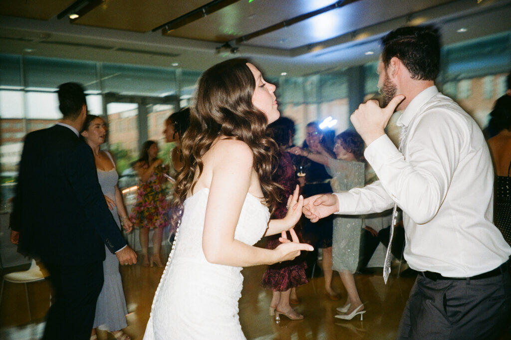 A bride and groom dance joyfully together in a modern, well-lit reception hall. Guests of various ages surround them, some dancing and others watching. The room features large windows and wooden floors, creating a warm and festive atmosphere.