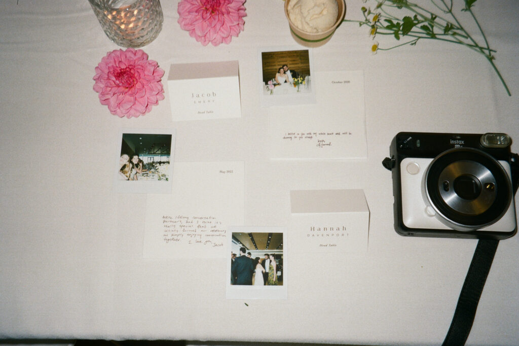 A table with pink flowers, a candle, and a camera. There are several cards and Polaroid photos, including names and dates, scattered around. One card reads Jacob and another Hannah. Cream in a small bowl is also visible.