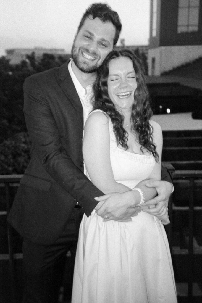 A black and white photo of a couple standing on a balcony. The man, in a suit, hugs the woman from behind. She is wearing a light dress. Both are smiling with eyes closed. Trees and buildings are visible in the background.