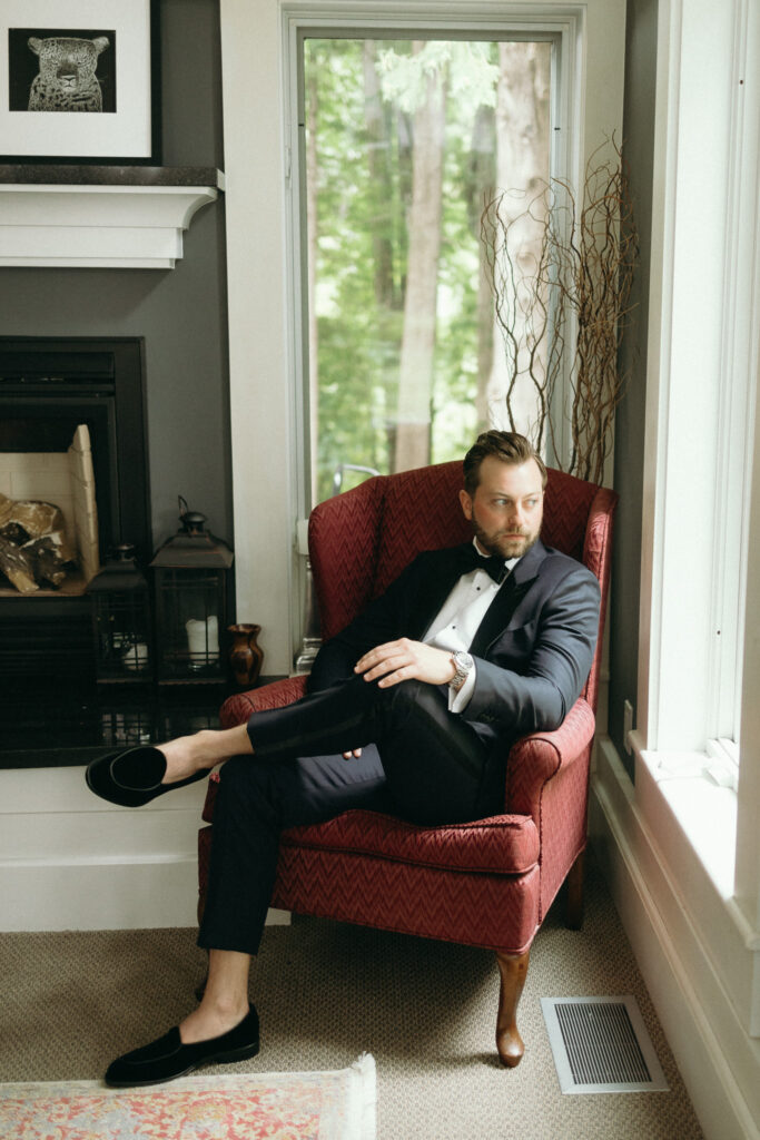 A man in a tuxedo sits casually in a red armchair with one leg crossed over the other. Hes in a room with large windows and a fireplace. The setting is elegant, with natural light coming through the windows.