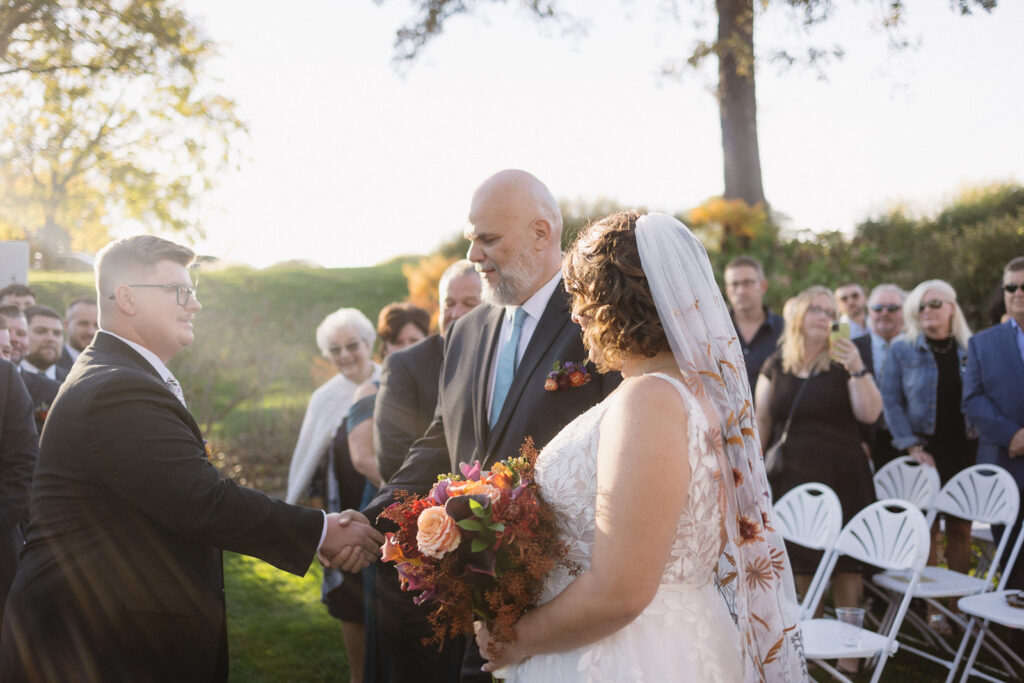 A groom shakes hands with a man giving away the bride at an outdoor wedding. The bride holds a bouquet of colorful flowers, wearing a veil. Guests are seated in white chairs under a sunny, tree-filled setting.