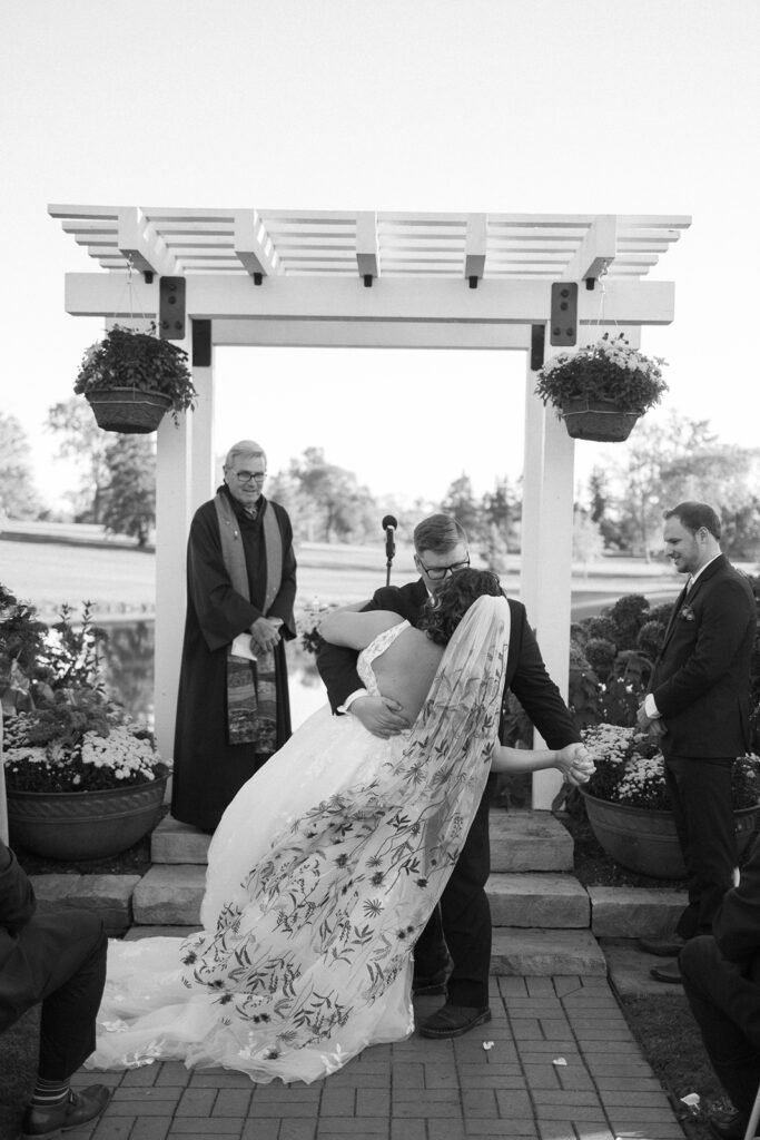 A couple shares a dip kiss under a white pergola during a wedding ceremony. The brides long veil with floral patterns is flowing, and a clergyman stands nearby. Guests are seated in the foreground, and the background shows a garden setting.