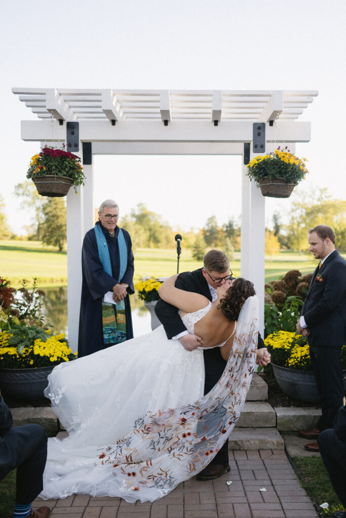 A couple shares a kiss during an outdoor wedding ceremony under a white pergola adorned with yellow flowers. The bride wears a flowing gown with an embroidered cape. A person in a robe stands nearby, with guests observing the moment.