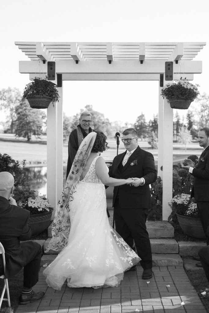 A couple holds hands and smiles during a wedding ceremony under a pergola, with an officiant behind them. Guests are seated nearby. The bride wears a long veil, and the groom is in a suit and sunglasses. The setting is outdoors with trees in the background.