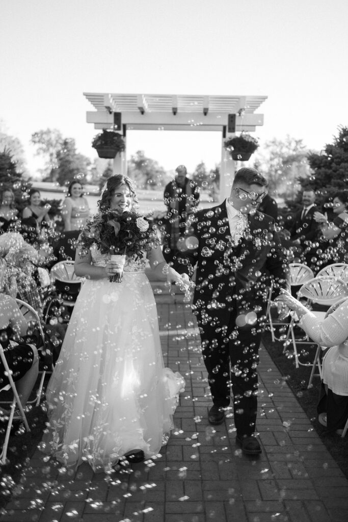 A newlywed couple walks down the aisle outdoors under a white pergola. The bride wears a flowing gown and holds a bouquet, while the groom is in a suit. Guests are seated on either side, and bubbles fill the air, creating a festive atmosphere.