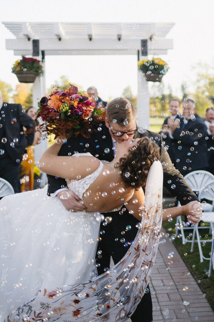 A bride and groom share a romantic kiss under a wooden arbor decorated with flowers. The groom holds the bride, who carries a colorful bouquet, as bubbles float around them. Guests in the background are seated, witnessing the joyous moment.