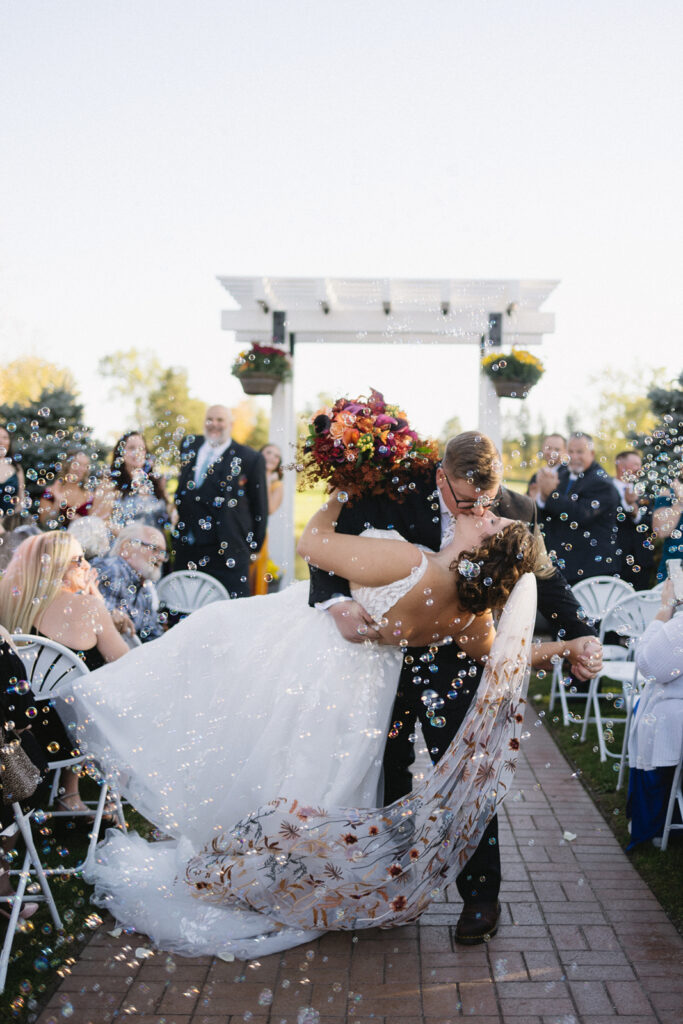 A bride in a white dress is being dipped and kissed by a groom in a black suit under a white pergola. She holds a bouquet of colorful flowers. Guests seated on white chairs clap and smile, while bubbles float around in the bright outdoor setting.