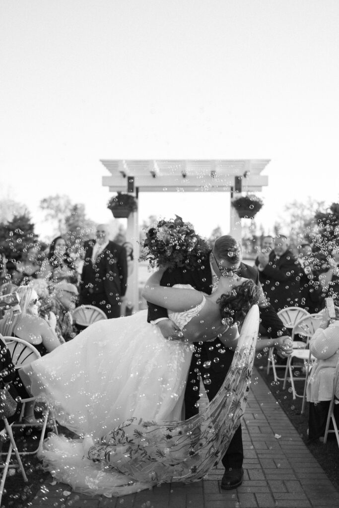 A couple kisses passionately as the groom dips the bride under a white pergola. The bride wears a flowing gown, and bubbles float around them. Guests seated on either side watch and smile. The scene is set outdoors in bright daylight.