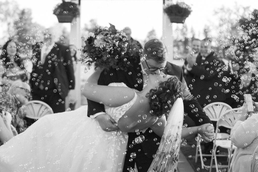 A couple kisses while the groom dips the bride during an outdoor wedding ceremony. Bubbles float around them, and guests are seated in the background. The scene is joyful and romantic in black and white.