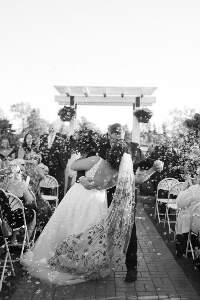 Bride and groom kissing at an outdoor wedding ceremony. The groom dips the bride, who wears a floral-patterned dress. Guests are seated on either side, blowing bubbles that float around the couple.