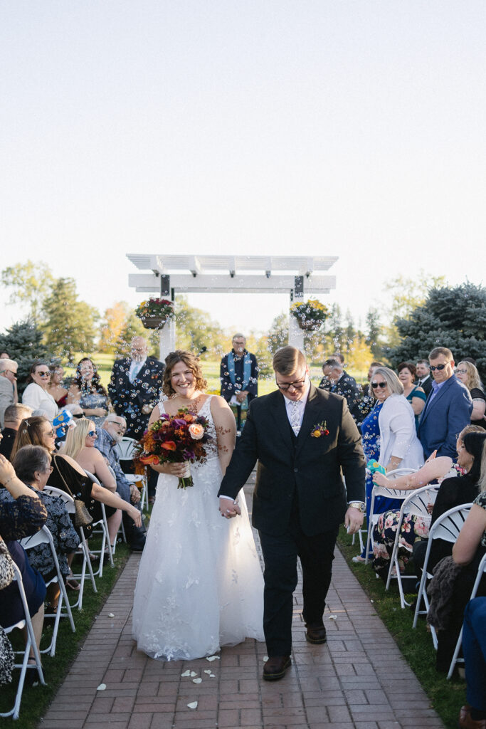 A wedding couple walks down an outdoor aisle. The bride wears a white gown and holds a bouquet with orange and red flowers. The groom is in a dark suit. Guests sit on either side, some smiling and taking photos. Trees and a white pergola are in the background.