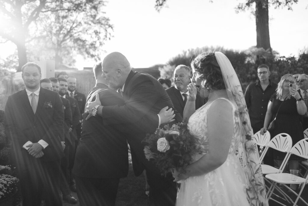 A black and white photo of a wedding ceremony outdoors. The bride, holding flowers, stands facing the aisle. A man hugs another man, likely the groom, while guests watch. Sunlight filters through the trees in the background.