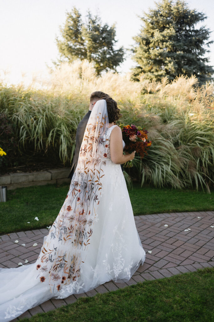 A bride and groom walk on a brick path. The bride wears a white dress with a long veil featuring floral embroidery, holding a bouquet of red and orange flowers. They are surrounded by tall grass and greenery.