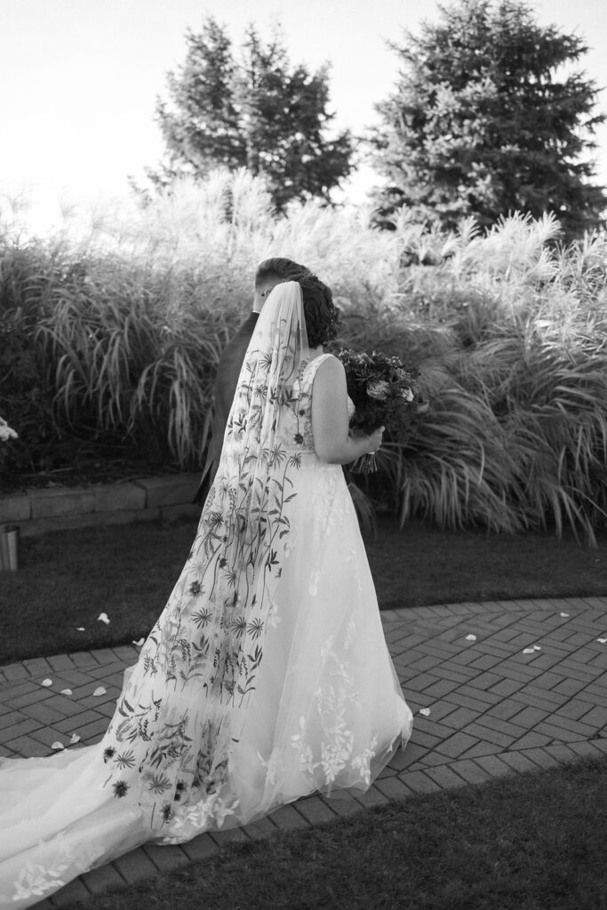 Bride in a floral-patterned veil holds a bouquet, walking with a groom in a garden setting. Theyre on a brick path surrounded by tall grasses and greenery. The scene is captured in black and white.