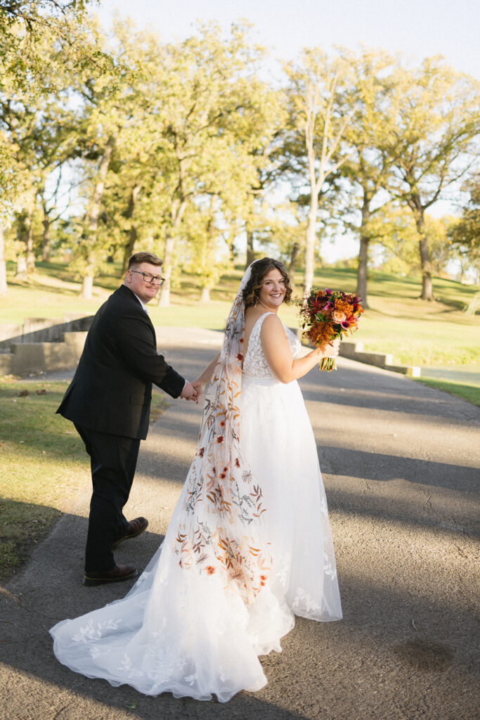 A bride and groom hold hands and walk on a sunlit path in a park. The bride wears a white dress with an embroidered veil and holds a bouquet of fall-colored flowers. The groom is in a black suit. Trees with autumn leaves surround them.