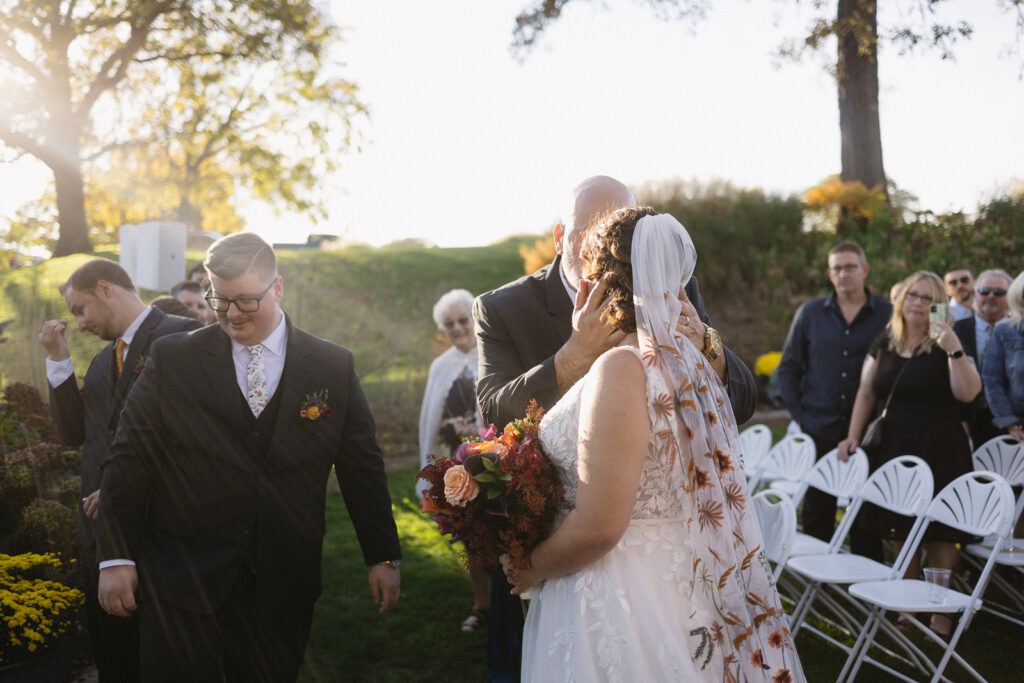 A groom embraces a bride outdoors during a sunny wedding ceremony. She holds a bouquet of flowers, and they stand on a grassy area. Guests are seated nearby, watching, with autumn trees in the background. The atmosphere is joyful and celebratory.