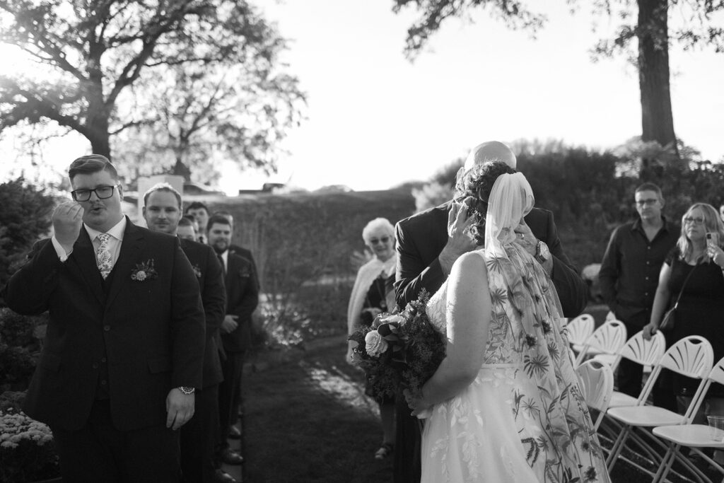 A bride in a floral dress and a veil hugs an elderly man during an outdoor wedding ceremony. Guests, some standing and some seated on white chairs, watch the moment. A man in a suit wipes a tear. Trees and sunlight create a serene backdrop. Black and white photo.