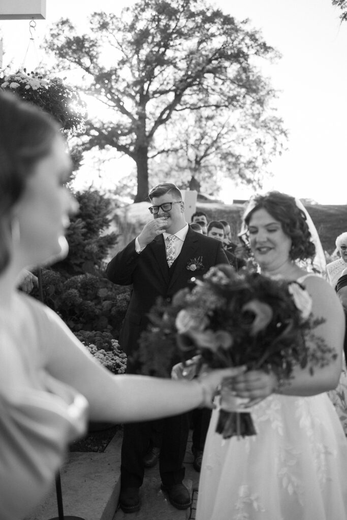 A groom in glasses wipes a tear while holding hands with the bride, who is carrying a bouquet. Another person stands in the foreground holding an item. There is a large tree and soft sunlight in the background. Black and white photo.