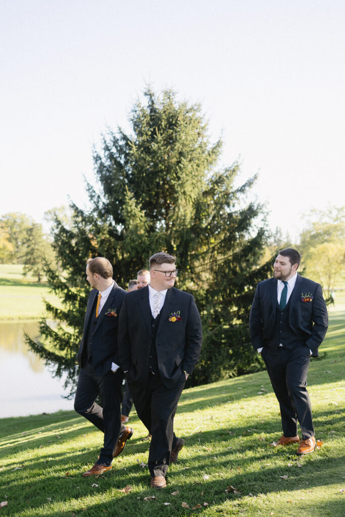 Three people in suits walk on a grassy field near a pond, with a tall evergreen tree in the background. They are dressed formally and appear to be in a cheerful conversation, enjoying a sunny day.