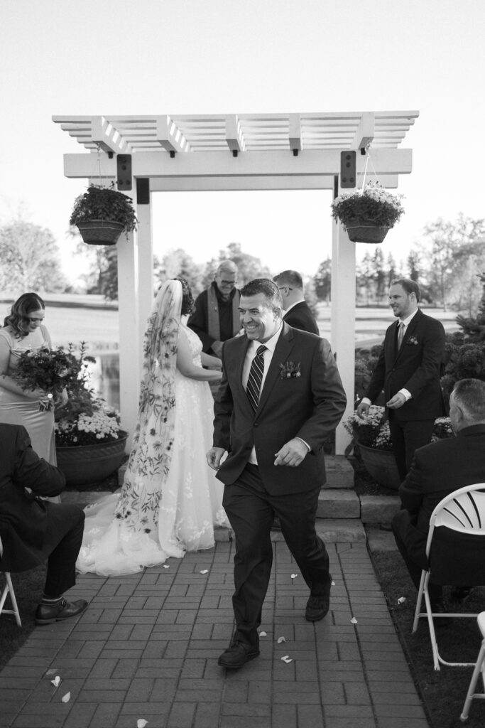 A joyful man in a suit walks down the aisle in an outdoor wedding ceremony. Behind him, a bride in a floral dress stands under a pergola adorned with hanging plants. Guests are seated on either side, and the scene is captured in black and white.