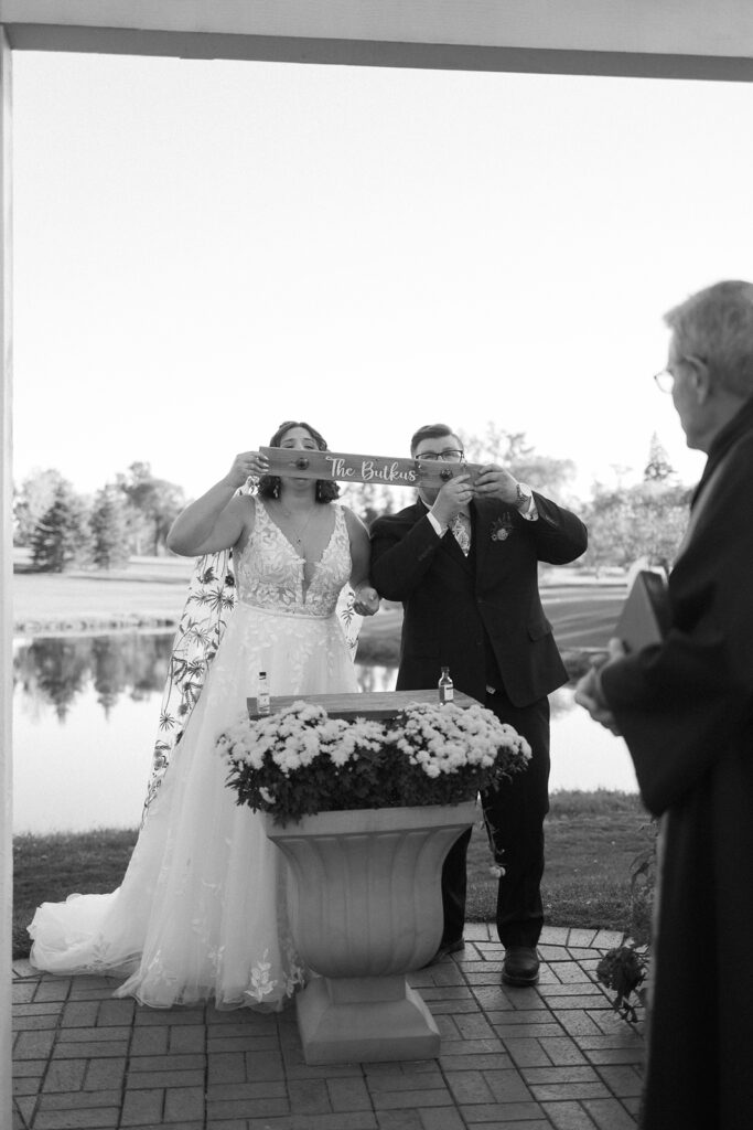 Bride and groom in wedding attire drink from personalized cups during an outdoor ceremony. A priest stands nearby holding a book. Flowers are arranged in a vase on a pedestal. A pond and trees are in the background. Black and white image.