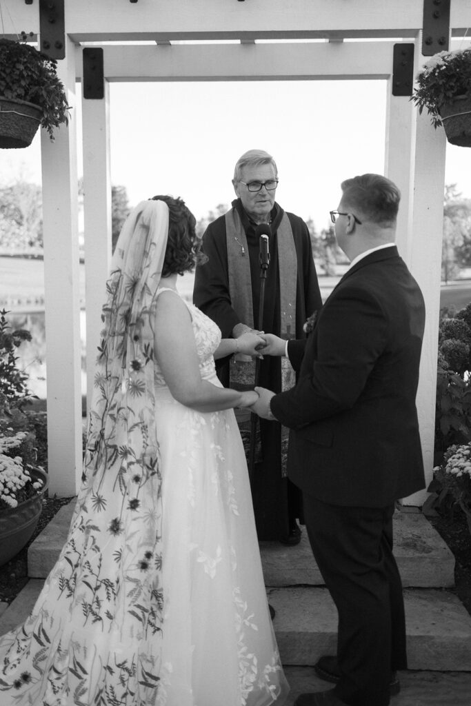 A bride and groom stand facing each other, holding hands during an outdoor wedding ceremony. An officiant wearing a robe stands between them, speaking into a microphone. The brides dress has floral embroidery, and they are beneath an archway.