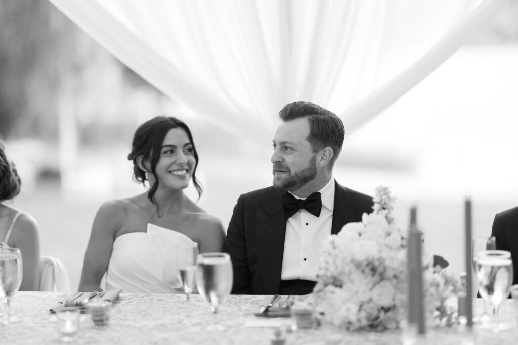 A black and white photo of a bride and groom seated at a wedding reception table. The bride is smiling at the groom, who is looking back at her. The table is decorated with flowers, candles, and glasses. Sheer curtains are visible in the background.