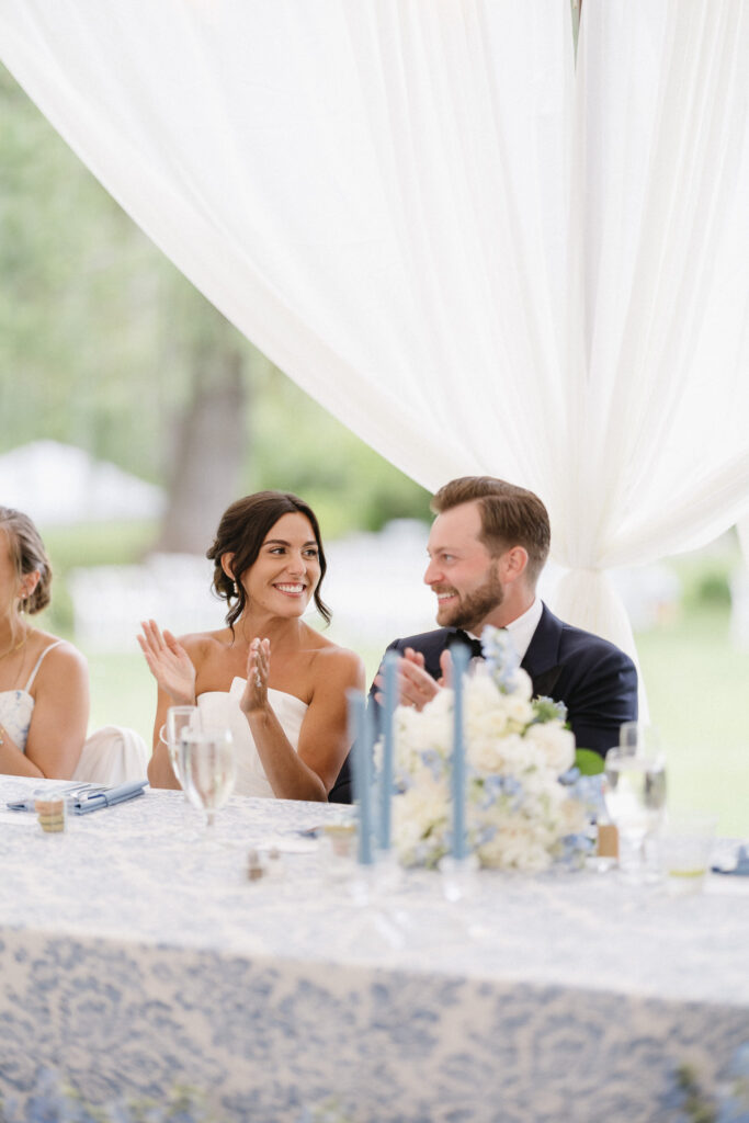 A bride and groom sit at a decorated table, smiling and clapping, under a white canopy. The table is adorned with blue and white flowers and lit candles. The background shows a blurred view of greenery.