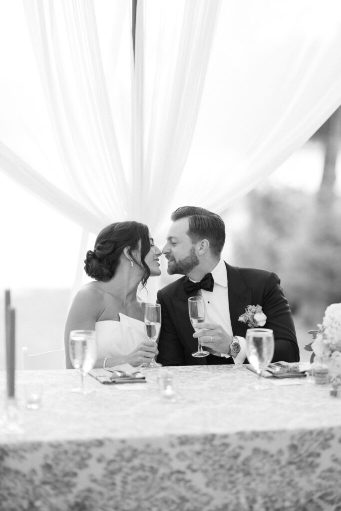 A bride and groom in formal attire share a romantic moment at a decorated table. Both hold champagne glasses and lean in closely. The setting is elegant with a draped backdrop and floral arrangements. The image is in black and white.