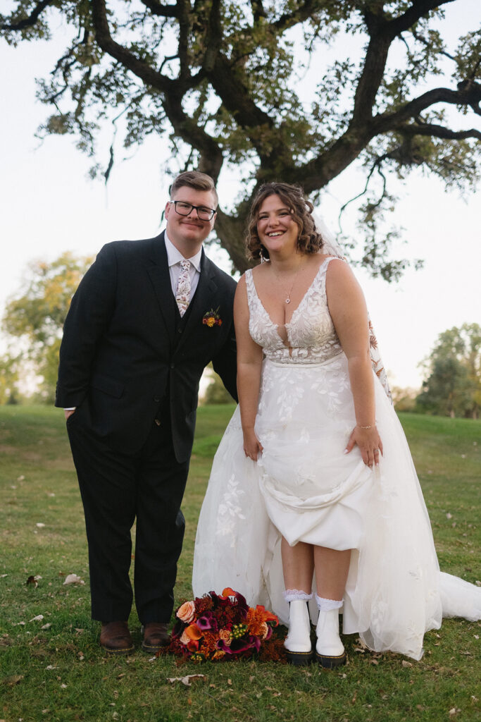 A newlywed couple stands outdoors under a tree. The bride, wearing a white gown and boots, leans forward smiling. The groom, in a dark suit with a boutonniere, stands beside her smiling. A bouquet of red and orange flowers lies on the grass.