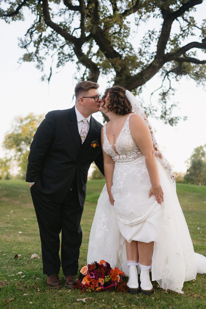 A bride and groom kiss outdoors under a large tree. The bride is in a white gown with a veil and the groom is in a dark suit with a tie. A bouquet of colorful flowers lies on the ground in front of them.