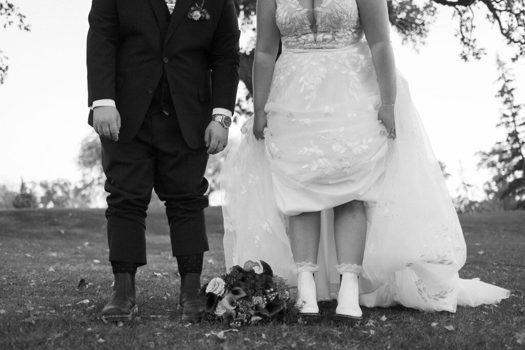 Black and white photo of a bride and groom standing on grass. The focus is on their legs and feet. The bride wears a gown with lace details and white boots. The groom is in a dark suit with floral socks and boots. A bouquet is on the ground between them.