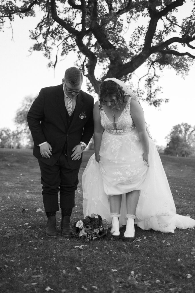 A black and white photo of a bride and groom bending forward to look down, possibly at a floral arrangement on the ground. They are outdoors, with trees and grass in the background. The bride is wearing a dress and the groom is in a suit.