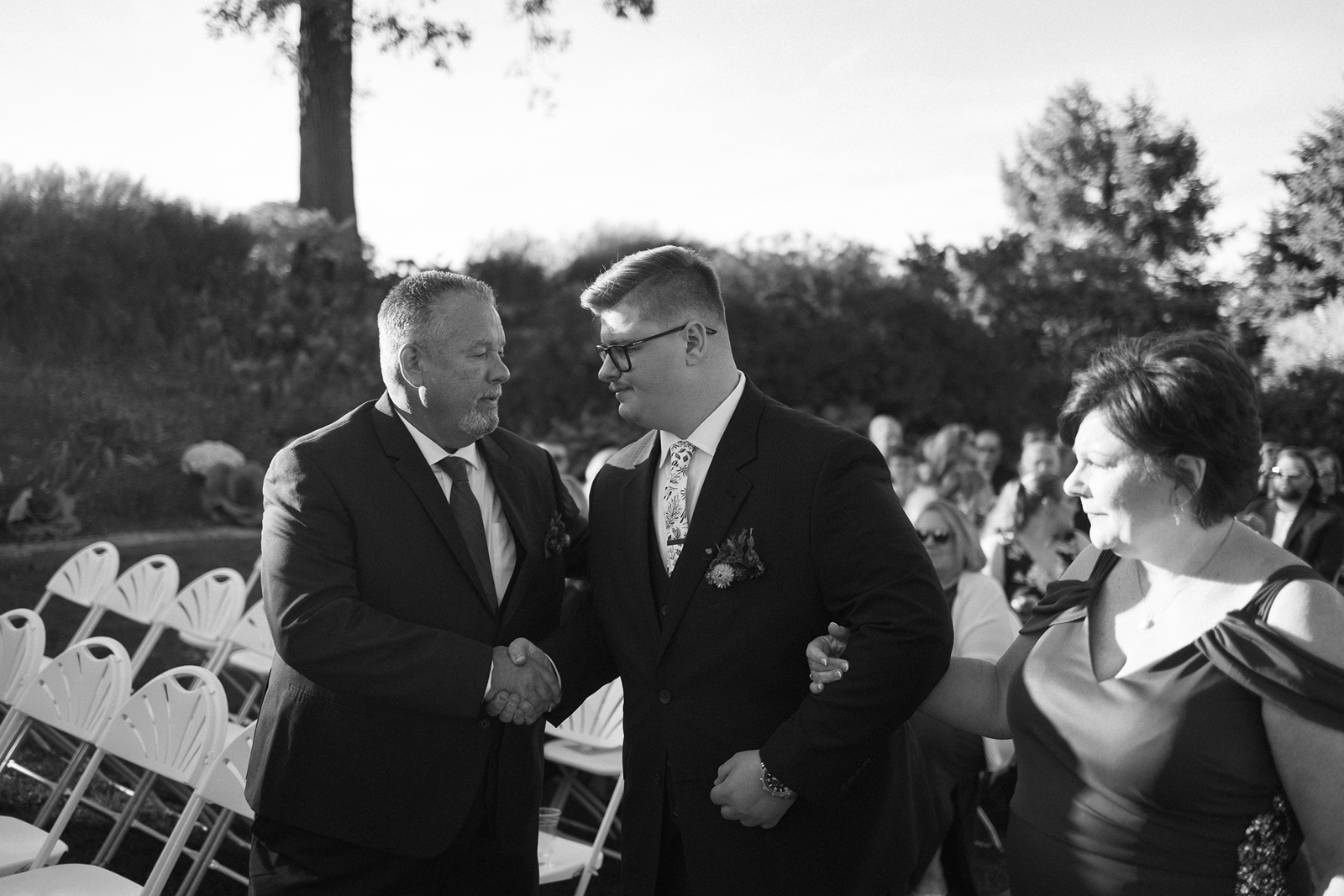 A black and white photo of a man and a younger man in suits, shaking hands. A woman in a dress stands beside them. They are outdoors, surrounded by white folding chairs and guests in the background. Trees and bushes are visible behind them.