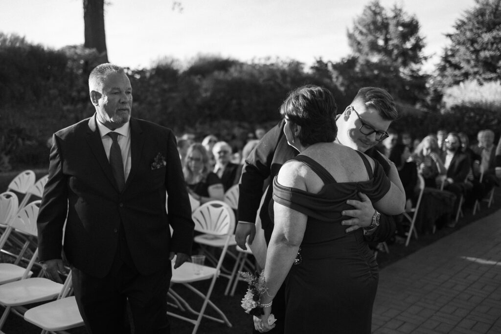 A black and white photo of a wedding ceremony outdoors. A person in a suit embraces a woman in a formal dress as they walk down the aisle. Guests are seated in the background, and a man follows closely. Trees and foliage line the area.