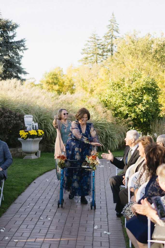 An elderly woman with a walker, decorated with flowers, walks down an outdoor garden aisle at a wedding ceremony. A seated man reaches out to her. Another woman follows behind. Guests are seated on either side, and trees are visible in the background.