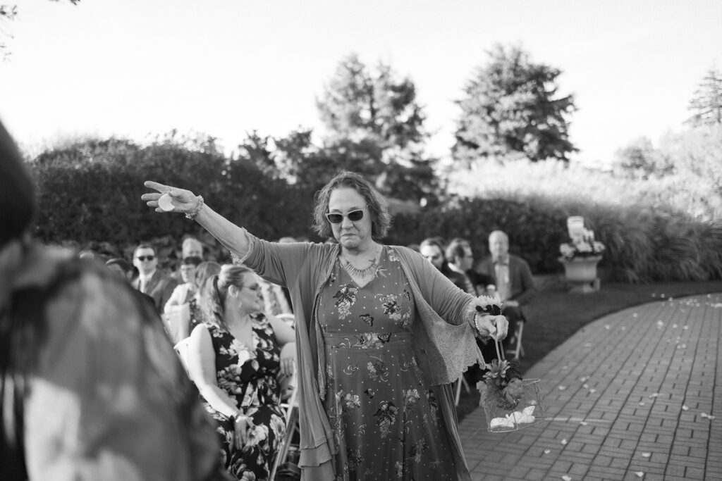 A woman in sunglasses and a floral dress walks down an outdoor aisle, holding a flower petal basket. She gestures with one arm extended. Seated guests watch as she proceeds, with trees and bushes visible in the background. Black and white image.