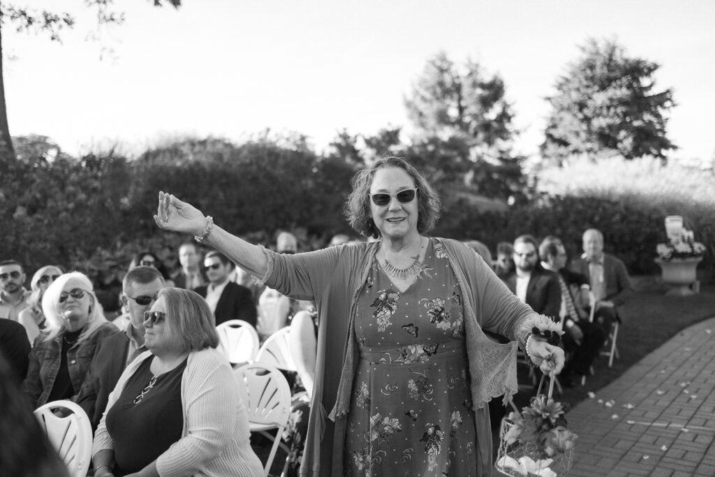 A woman in a floral dress and sunglasses gestures joyfully at a wedding. She stands amidst seated guests on white chairs in an outdoor setting with trees and bushes in the background. The mood is festive and lively.