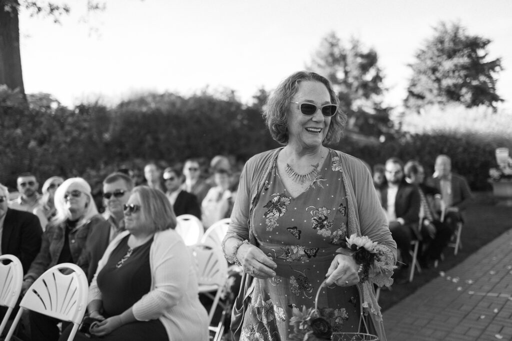 A smiling woman in sunglasses and floral dress walks up an aisle at an outdoor event. She holds a basket with flowers. Seated guests in sunglasses watch her. Its a sunny day with trees in the background. Black and white image.