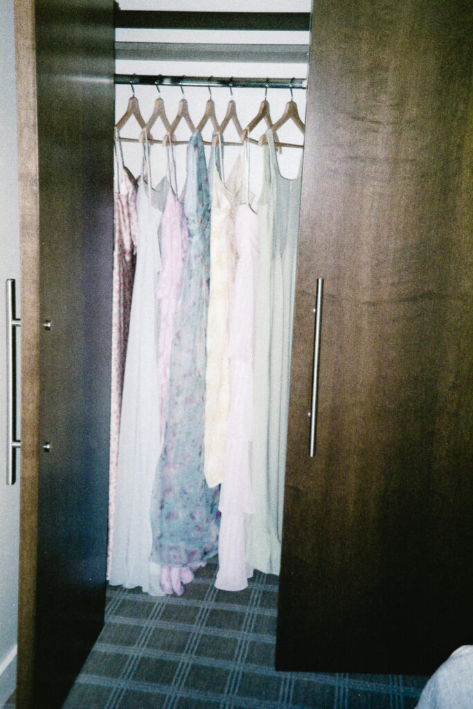 An open wardrobe with wooden doors reveals several pastel and floral dresses hanging inside on wooden hangers. The floor is carpeted in a gray pattern.