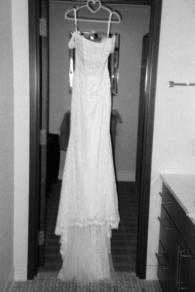 Black and white image of an elegant lace wedding dress hanging on a door, with a heart-shaped hanger. The dress has an off-the-shoulder neckline and a flowing train, set in a room with a mirror and a bathroom counter visible.