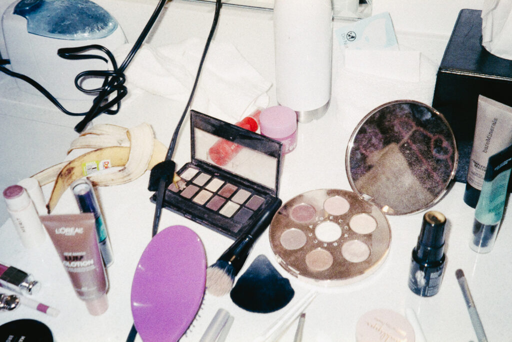 A cluttered vanity with makeup items, including eyeshadow palettes, lipsticks, brushes, and facial products. A banana peel is visible among the cosmetics. Items are scattered across a white surface.