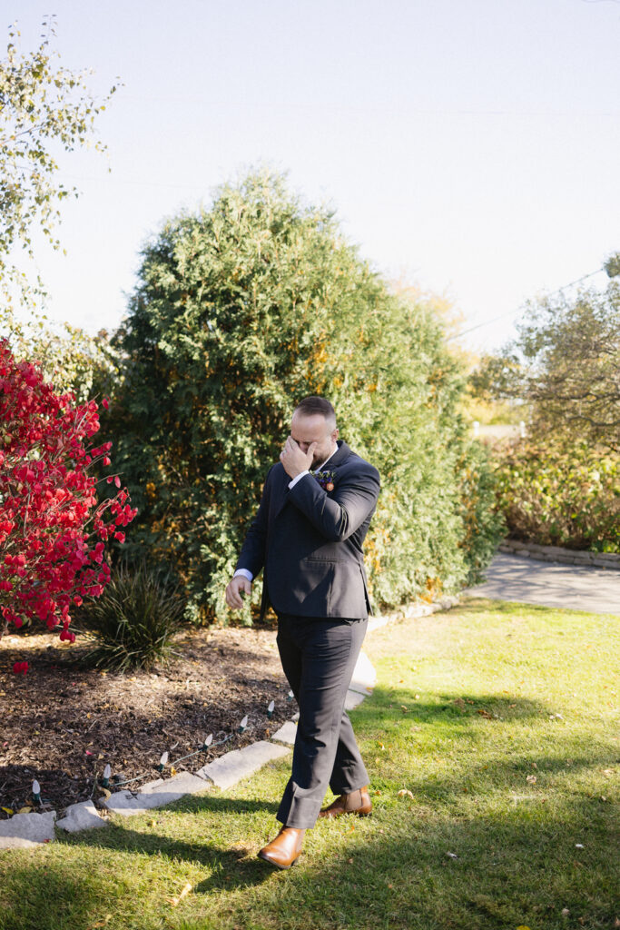 A man in a formal suit walks across a grassy area, covering his mouth with his hand. He is near a garden with green bushes and a red-leaved shrub. The sky is clear and sunny.