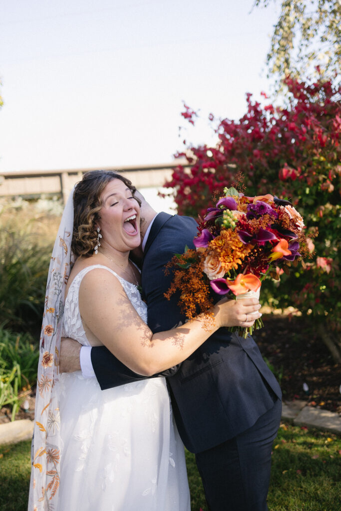 A joyful bride laughing as she hugs the groom outdoors. She holds a vibrant bouquet with orange, purple, and red flowers, while wearing a white dress and veil. The groom is in a dark suit. The background features greenery and a blooming bush.