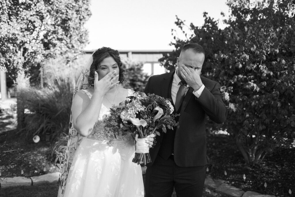 A bride and groom stand outdoors, each wiping tears from their eyes. The bride holds a bouquet and wears a floral gown. The groom is in a suit. They are surrounded by lush greenery, with a building visible in the background.