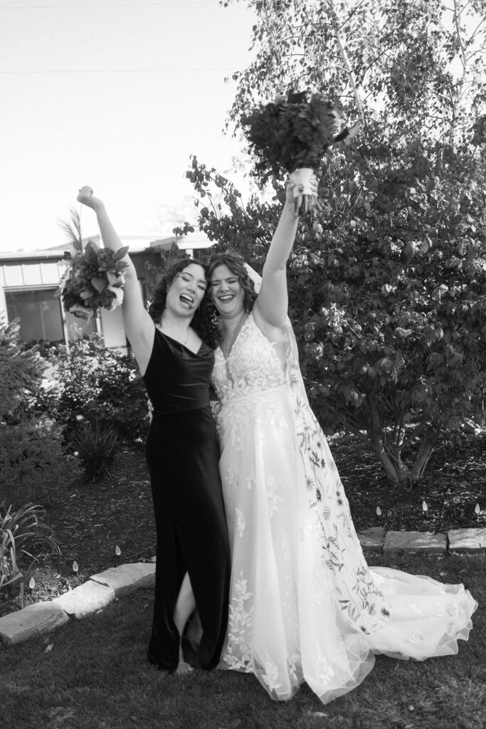 Two women celebrate outdoors, both holding bouquets and raising their arms joyfully. One wears a long black dress, the other a floral wedding dress. They stand on a stone path surrounded by greenery.