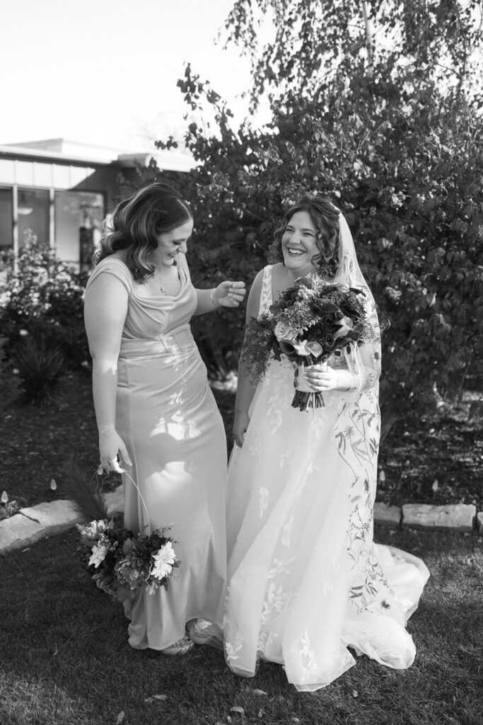 Two women in a garden, one in a wedding dress holding a bouquet, smiling and laughing. The other woman is in a bridesmaid dress, holding a smaller bouquet. Theyre surrounded by greenery on a sunny day. Black and white photo.