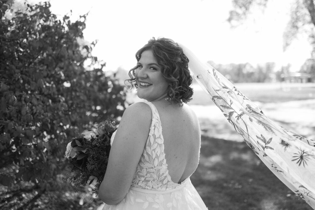 A bride stands outdoors, smiling over her shoulder. She wears a gown with floral embroidery and a long veil flowing behind her, adorned with delicate patterns. Sunlight filters through the trees, casting a serene glow.