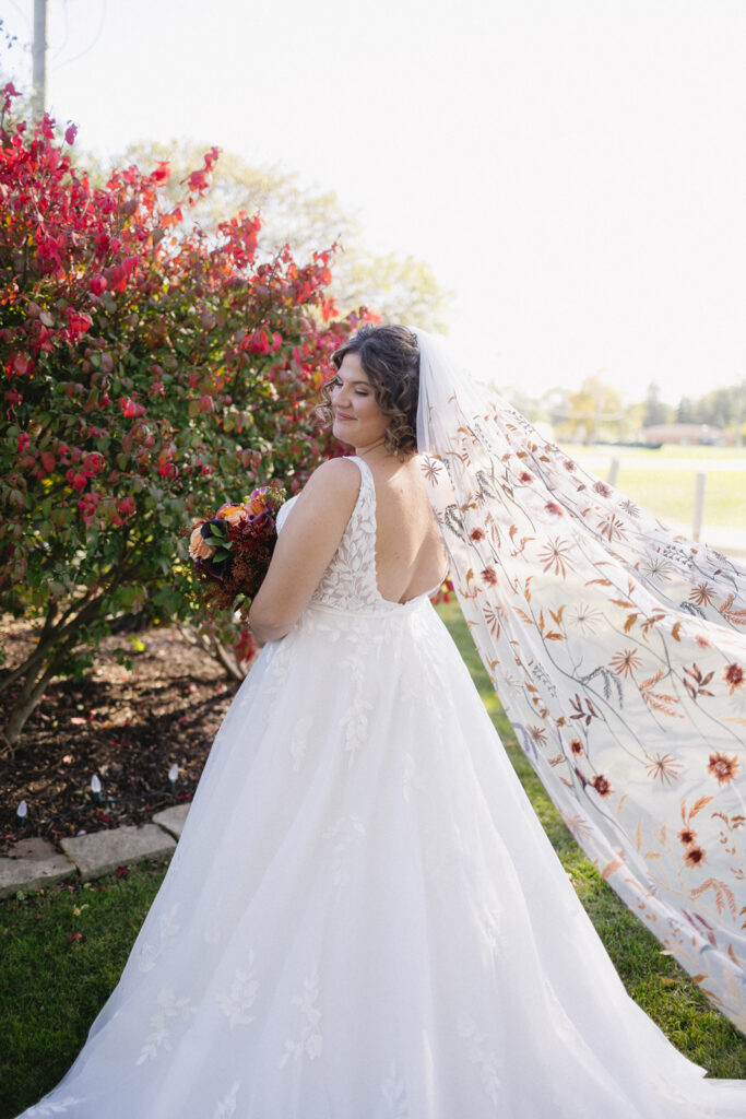 Bride in a white wedding gown stands outdoors, facing slightly away. Her long veil is adorned with floral patterns and flows behind her. She holds a bouquet of red flowers, with a lush garden and bright sky as the backdrop.