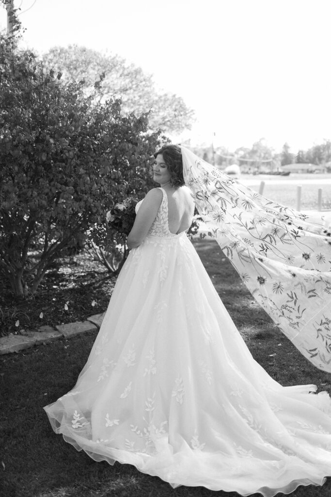 A bride stands outdoors in a wedding dress with floral embroidery, holding a bouquet. Her veil, also with floral patterns, flows in the breeze. She smiles while looking over her shoulder, with trees and a body of water in the background.